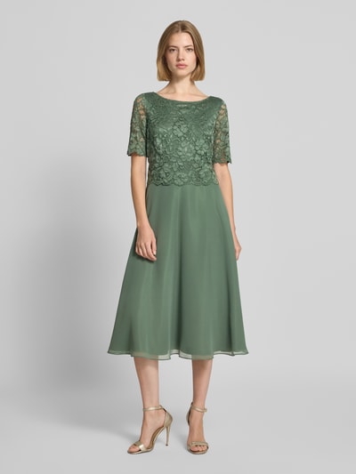 Frauenmodel trägt Tailliertes Cocktailkleid mit Spitzenbesatz von Vera Mont in Salbeigrün, florale Spitze, fließender Rock, Ganzkörperansicht von vorne. Damen Cocktailkleid von Vera Mont.