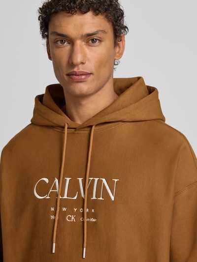 Männermodel draagt Hoodie mit Label-Print und Känguru-Tasche van CK Calvin Klein in bruin, katoen-elastaan, relaxed fit, Nahaufnahme Oberkörper.