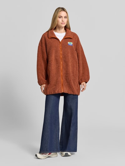 Frauenmodel trägt Oversized Jacke mit Stehkragen Modell 'Hoktown' von American Vintage aus braunem Fleece mit Reißverschluss, Logo-Patch, Ganzkörperansicht von vorne.