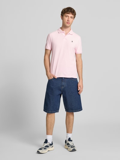 Męski model nosi Modern Fit Poloshirt mit Logo-Stitching od REVIEW, bawełniany miks, krótka listwa guzikowa, kolor jasnoróżowy, granatowe jeansowe szorty, pełny widok z przodu.