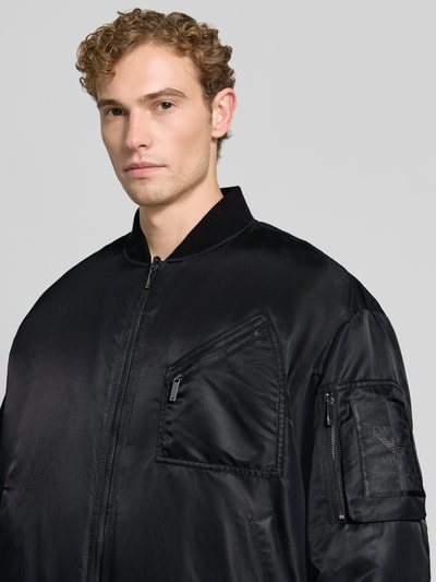 Männermodel trägt schwarze Bomberjacke mit Stretch-Anteil von Emporio Armani, Satinoptik, Zipperdetails, Nahaufnahme Oberkörper.
