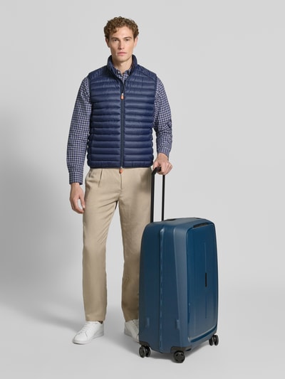 Männermodel trägt kariertes Hemd, blaue Steppweste, beiger Hose, hält Koffer mit Logo-Detail Modell 'Spinner' von Samsonite, Ganzkörperansicht von vorne.
