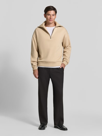 Męski model nosi Regular Fit Sweatshirt aus Baumwoll-Mix mit Troyer-Kragen od DRYKORN, mieszanka bawełny, krój regular fit, kołnierz zapinany, wyhaftowane logo, Długość tyłu w rozmiarze M: 64 cm, Długość rękawów w rozmiarze M: 57 cm, beżowa góra, czarne spodnie, gładkie faktury, Ganzkörperansicht von vorne.