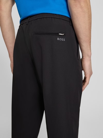 Männermodel trägt Tapered Fit Anzughose aus bügelleichtem Material Modell 'T_CONVERT' von BOSS Green, zwarte effen kleuren, regular fit, tunnelkoord, kort, persplooien, sneldrogend, geïntegreerd kaartetui, zij-steekzakken, 4-weg stretch materiaalverwerking, detailaufnahme achterkant.