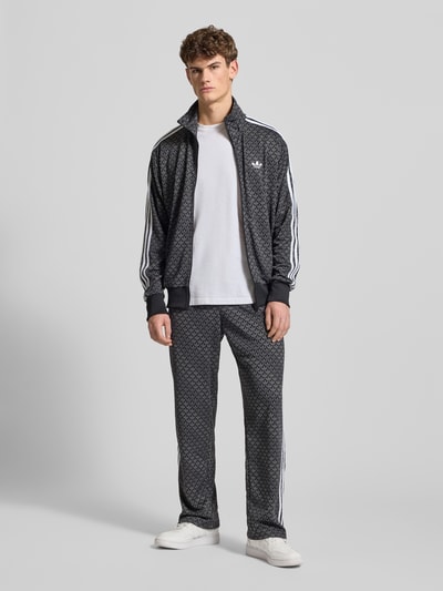 Männermodel trägt Sweatjacke mit Stehkragen von adidas Originals in Schwarz mit weißem Label-Stitching, Lockerer-Schnitt, Reißverschluss, Reißverschlusstaschen, gerippte Abschlüsse, Rückenlänge 72 cm, Ärmellänge 65 cm, Ganzkörperansicht von vorne.