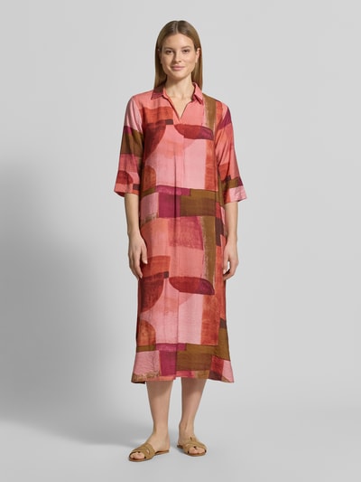 Frauenmodel trägt Relaxed Fit sukienka midi z mieszanki wiskozy model 'Catania' od Soyaconcept w odcieniach różu, czerwieni i brązu z geometrycznym wzorem, wykładany kołnierz, rękaw o dł. 1/2, dekolt w serek, długość tyłu w rozmiarze S: 130 cm, szerokość w rozmiarze S: 58 cm, długość rękawów w rozmiarze S: 41 cm, Ganzkörperansicht von vorne.