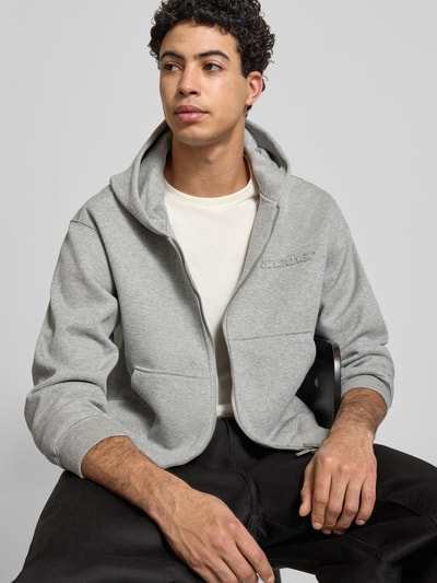 Männermodel trägt Sweatjacke mit Kapuze von ANOTHER COTTON LAB in Hellgrau, lockerer Schnitt, sichtbare Taschen, Nahaufnahme Oberkörper. Baumwoll-Mix mit Stretch-Anteil, Reißverschluss und Label-Detail.