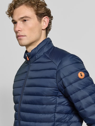 Männermodel trägt dunkelblaue Steppjacke mit Zweiwege-Reißverschluss Modell 'ALEXANDER' von Save The Duck, Nahaufnahme Oberkörper, Slim Fit, Stehkragen, sichtbares Markenlogo, glatte Oberfläche