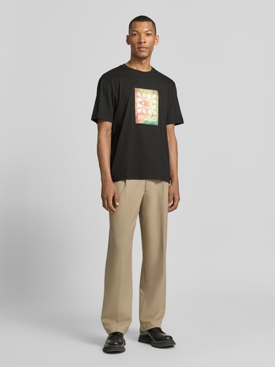 Männermodel trägt Regular Fit T-Shirt aus reine Baumwolle Modell 'DAMORU' von HUGO in Schwarz mit buntem Foto-Print, Ganzkörperansicht von vorne.