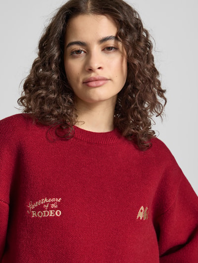 Frauenmodel trägt Relaxed Fit Pullover mit Logo-Stitching von ANOTHER COTTON LAB in Rot, Strick-Optik, Nahaufnahme Oberkörper.