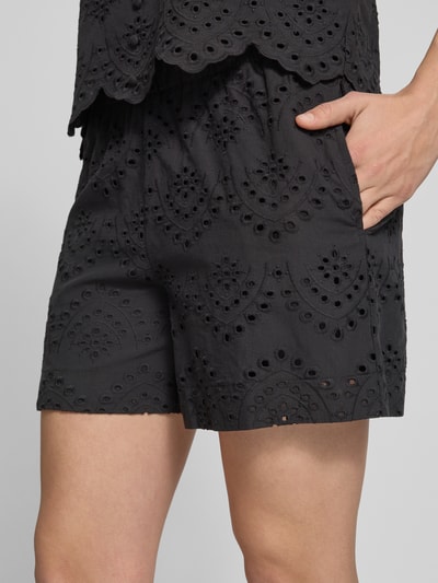 Frauenmodel trägt Loose Fit Bermudas mit Lochstickerei Modell 'VILDE' von Pieces, schwarze Baumwolle, seitliche Taschen, Nahaufnahme Oberschenkel.