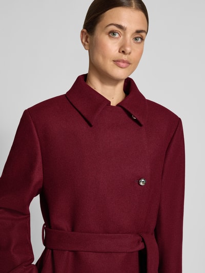 Frauenmodel trägt Mantel mit Bindegürtel Modell 'Melm' von HUGO in Unifarbe Bordeaux mit Zierknöpfen und Teilungsnähten, Nahaufnahme Oberkörper.