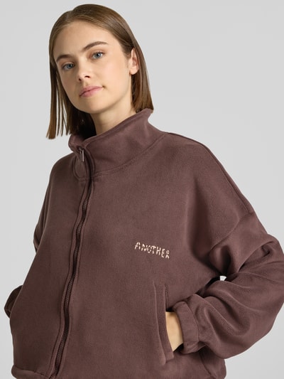 Modelka kobieca nosi Cropped Fleecejacke mit Label-Detail von ANOTHER COTTON LAB, brązowy materiał z widoczną teksturą, zamek, stójka, zbliżenie na górę ciała
