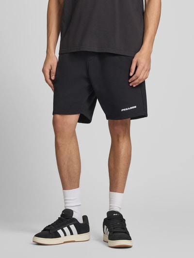 Männermodel trägt Shorts mit elastischem Bund und Logo-Stitching von Pegador aus Baumwoll-Mix, Loose-Fit, mit Eingrifftaschen, Nahaufnahme Oberkörper.