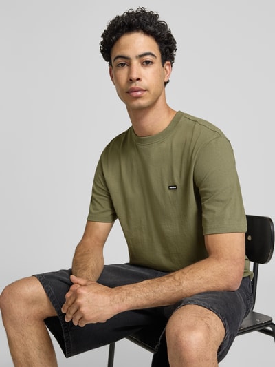 Männermodel trägt T-Shirt mit Label-Patch und Rundhalsausschnitt von REVIEW, Boxy Fit, reine Baumwolle, Olivgrün, Nahaufnahme Oberkörper.