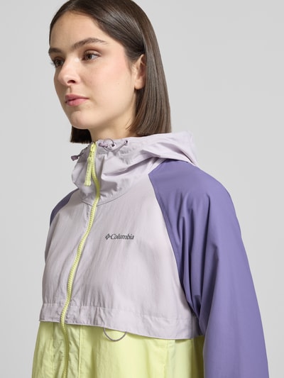 Frauenmodel trägt Windbreaker mit Rückenbelüftungssystem und Kapuze von COLUMBIA in zartem Violett und Gelb, Logo-Print, Nahaufnahme Oberkörper. Kontext: Lockerer-Schnitt, Kapuze mit Kordelzug, Omni-Shield™ Technologie, wasser- und schmutzabweisend, Kinnschutz, Rückenbelüftungssystem, Eingrifftaschen mit Reißverschluss, elastische Bündchen, Saum mit verstellbarem Kordelzug, Rückenlänge 59 cm, Schulterbreite 45 cm, Brustweite 51 cm, Ärmellänge 57 cm.