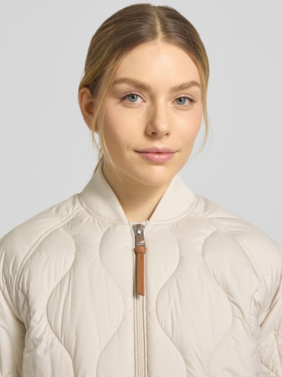 Frauenmodel trägt Steppmantel mit Ärmeltasche Modell 'AVON' von Beaumont Amsterdam in Beige, gesteppte Struktur, Nahaufnahme Oberkörper, gerade Passform, Stehkragen, Zweiwege-Reißverschluss.