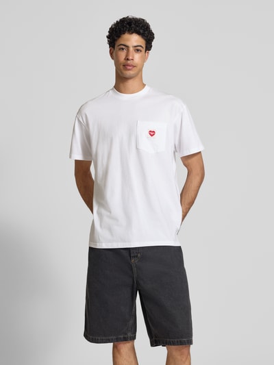 Männermodel trägt T-Shirt mit Brusttasche und Label-Patch von Carhartt Work In Progress in Weiß, gerade Passform, Ganzkörperansicht von vorne.
