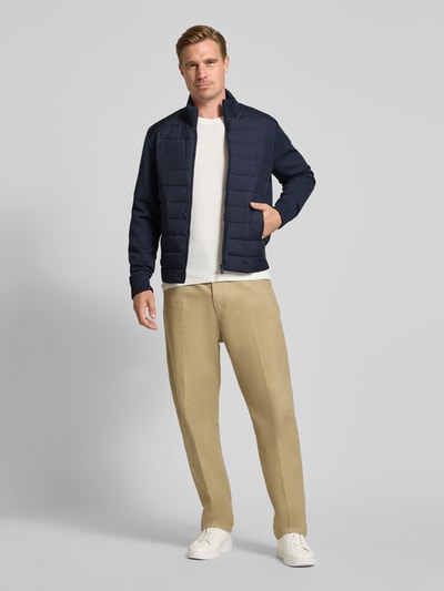 Männermodel trägt Steppjacke mit geripptem Stehkragen Modell 'SEDUM' von Save The Duck in Dunkelblau, helle Hose, glatte Texturen, Ganzkörperansicht von vorne. Gerader Schnitt, Reißverschluss, Ärmel im Strick-Design, Label-Patch, seitliche Reißverschlusstaschen.