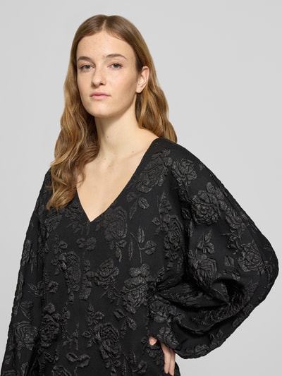 Vrouwenmodel draagt Minikleid mit V-Ausschnitt van Gina Tricot, zwart, losse fit, V-hals, sierbies, elastische mouwuiteinden, detailopname bovenlichaam.