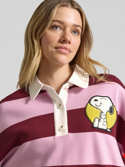 Modelka kobieca nosi Oversized Poloshirt aus Viskose-Mix Modell 'JICE STRIPED EMBO' od Essentiel Antwerp, wielokolorowe, w pasy, z motywem aplikacji Snoopy, kołnierzyk polo, drobny dzianinowy materiał, detal tekstowy, krótka zapięcie na guziki, rękaw 3/4, wykończenia prążkowane. Zbliżenie na górną część ciała.