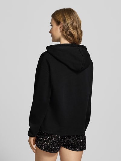 Modelka kobieca ma na sobie Regular Fit Hoodie aus Baumwoll-Mix Modell 'CHILLI' od Pieces, kolor czarny, detal z logo, kaptur z troczkiem, długie rękawy, ściągacze, gładka tekstura, widok całej sylwetki z tyłu.