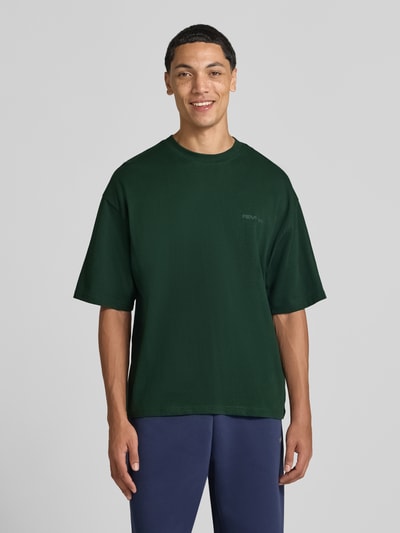 Męski model nosi Oversized T-Shirt aus reiner Baumwolle von REVIEW, ciemnozielony, bawełna, okrągły dekolt, nadruk z logo, długość tyłu w rozmiarze M: 66 cm, krój oversized, widok całej sylwetki od przodu.