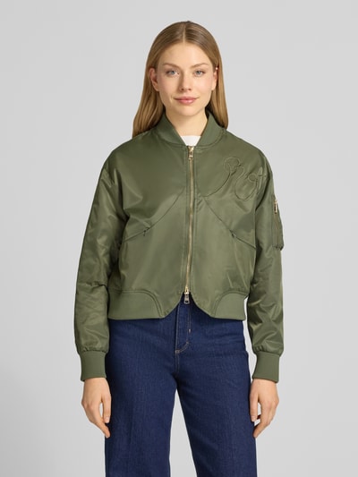 Frauenmodel trägt Bomberjacke mit Zweiwege-Reißverschluss von Original Bombers in Olivgrün mit Stehkragen und Stitching, Ganzkörperansicht von vorne.