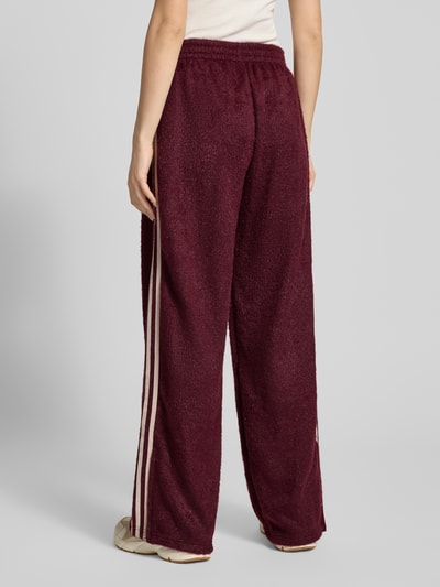 Vrouwenmodel draagt Sweatpants met elastische band van adidas Originals in bordeauxrood met glittereffect, labelstitching en ritszakken, los model, binnenbeenlengte maat S 81 cm. Ganzkörperansicht Rückansicht.