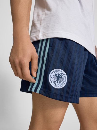 Mężczyzna nosi Loose Fit Trikot-Shorts mit DFB-Applikation od adidas Originals, pasiasty wzór, logo, elastyczny pas, tkanina żakardowa, detal z boku; zbliżenie na biodra.