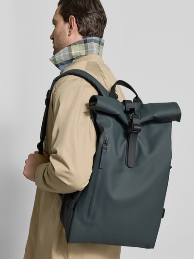 Männermodel trägt Rolltop Rucksack mit Reißverschlusstasche von Rains in Dunkelgrün mit Karabinerhaken, Tragehenkel, verstellbare Träger, Ganzkörperansicht Rückansicht. Breite 31 cm, Länge 61 cm, Tiefe 15 cm, Schulterriemen 74 cm.