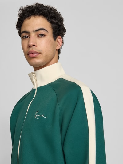 Männermodel trägt Regular Fit Sweatjacke mit Stehkragen von KARL KANI in Grün mit Logo-Stitching, Reißverschluss, Elastischer-Material-Mix, Eingrifftaschen, elastische Abschlüsse, Nahaufnahme Oberkörper.