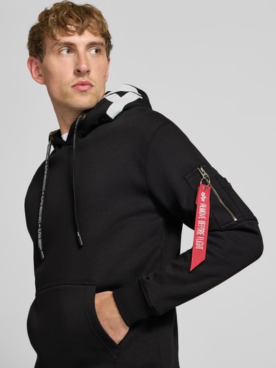Männermodel trägt schwarzen Regular Fit Hoodie aus Baumwoll-Mix mit Label-Patch von Alpha Industries, Nahaufnahme Oberkörper. Herren Hoodie mit Ärmel-Tasche, Känguru-Tasche, Kapuze mit Tunnelzug, Rückenlänge 67 cm, Ärmellänge 63 cm.