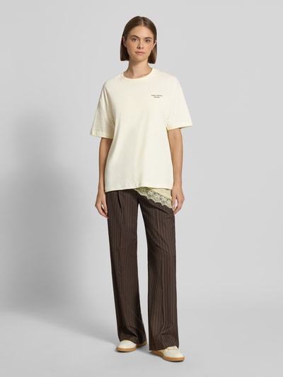Frauenmodel trägt Relaxed Fit T-Shirt aus reiner Baumwolle von Marc O'Polo DENIM in Creme mit glatter Struktur, kombiniert mit brauner Hose, Ganzkörperansicht von vorne.