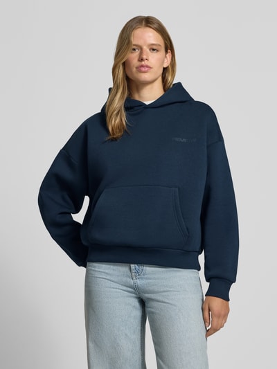 Frauenmodel trägt Kapuzenpullover aus Baumwoll-Mix von REVIEW in Dunkelblau mit Känguru-Tasche, lockerer Passform und gerippten Bündchen, Ganzkörperansicht von vorne.