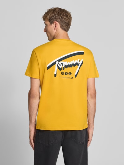 Männermodel trägt Regular Fit T-Shirt aus reiner Baumwolle von Tommy Jeans in Gelb mit großem weißen und schwarzen Print, Ganzkörperansicht Rückansicht.