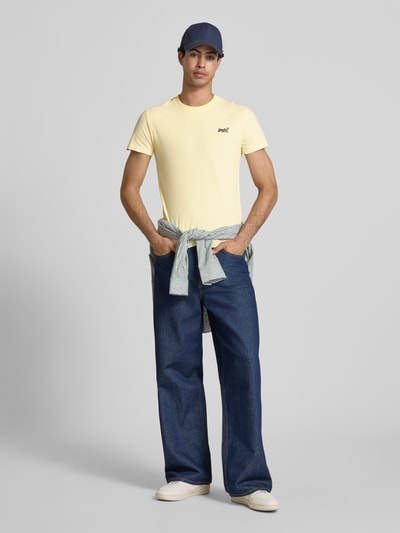 Männermodel trägt Regular Fit T-Shirt mit Logo-Stitching von Superdry in Hellgelb, kombiniert mit Jeans, Rundhalsausschnitt, Ganzkörperansicht von vorne.