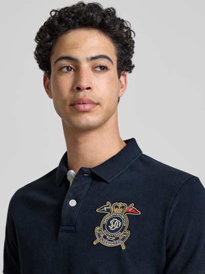 Männermodel trägt Regular Fit Poloshirt mit Label-Patches von Superdry aus Reine Baumwolle mit Polokragen und Knopfleiste, Nahaufnahme Oberkörper. Rückenlänge bei Größe M: 68 cm.