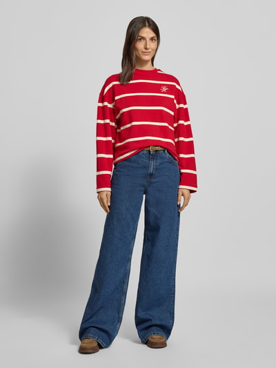 Frauenmodel trägt Relaxed Fit Sweatshirt aus Baumwoll-Mix von Tommy Hilfiger in Rot mit weißen Streifen und Logo-Stitching, Ganzkörperansicht von vorne.