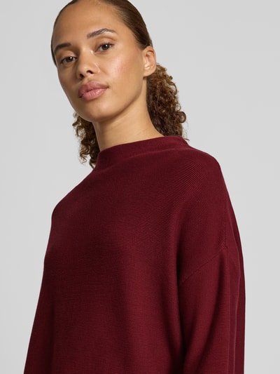 Frauenmodel trägt Loose Fit Pullover aus reiner Baumwolle Modell 'MERINAA' von ARMEDANGELS in Dunkelrot, feine Strickstruktur, Nahaufnahme Oberkörper.