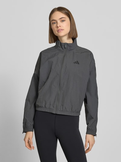 Modelka prezentuje Regular Fit Jacke mit Stehkragen od adidas Sportswear, szara, logo-print, zamek, elastyczne wykończenia, długość tyłu w rozmiarze S: 54 cm, szerokość ramion w rozmiarze S: 64 cm, szerokość klatki piersiowej w rozmiarze S: 64 cm, długość rękawa w rozmiarze S: 48 cm, całkowity widok z przodu
