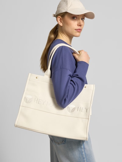 Frauenmodel trägt cremefarbene Handtasche mit Logo-Stitching Modell 'Signature' von Hey Marly, strukturierte Oberfläche, Ganzkörperansicht von vorne. Hauptfach mit Druckknopfverschluss, Reißverschlussfach innen, breiter Schulterriemen, Maße 42x31x15 cm, Riemenlänge 55 cm.