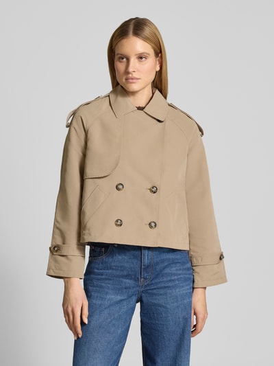 Kobiece modelka nosi Boxy Fit Trenchcoat mit doppelreihiger Knopfleiste Modell 'CHLOE' od Vero Moda, beżowy, gładki, długi rękaw raglanowy, regulowane mankiety, ukośne kieszenie, kołnierz wykładany, detal gun flap i kontrafałda, widok całej sylwetki z przodu.