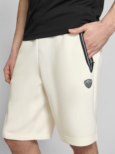 Męski model nosi Shorts mit elastischem Bund od EA7 Emporio Armani, kolor kremowy, krój regular fit, materiał z domieszką wiskozy, szerokość w rozmiarze M: 41 cm, długość nogawki wewnętrznej w rozmiarze M: 28 cm, zbliżenie na górną część ciała.
