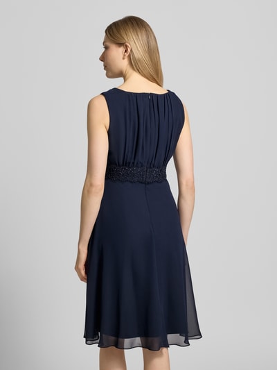 Frauenmodel trägt Knielanges Cocktailkleid mit Taillenband von Christian Berg Cocktail aus dunkelblauem Chiffon, Taillierter Schnitt, mit Rundhalsausschnitt und Drapierungen, Rückenlänge bei Größe 36: 103 cm, Brustweite bei Größe 36: 36 cm, Ganzkörperansicht Rückansicht.