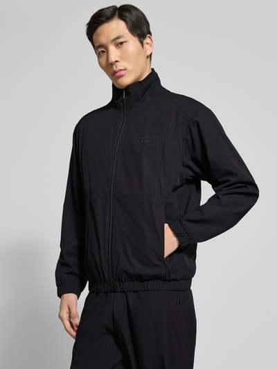 Mannelijk model draagt Regular Fit Trainingsanzug met Label-Detail Modell LW_PS Spirit70 TT - PORSCHE X BOSS van BOSS Green in effen zwart piqué, labelopschrift en label in reliëf, ritssluiting, tunnelkoord, elastische boorden, zijritszakken, tweedelig, look: effen, regular fit pasvorm, opstaande kraag, lange mouwen, materiaalverwerking: piqué, nahaufnahme bovenlichaam.