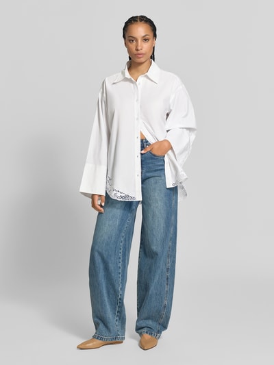 Frauenmodel trägt Oversized Hemdbluse mit Spitzenbesatz von Officine Générale in Weiß aus reiner Baumwolle mit Hemdblusenkragen und langärmeligem Design, lockerer Schnitt, kombiniert mit blauer Jeans und beigen Schuhen, Ganzkörperansicht von vorne.