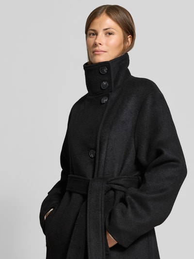 Frauenmodel trägt Mantel mit Baumwoll-Anteil von MYON in Schwarz, glatte Textur, Reverskragen, Taillienband, Knopfleiste, Nahaufnahme Oberkörper.