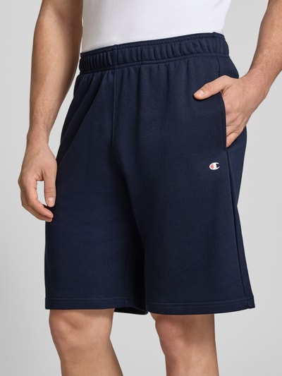 Männermodel trägt Shorts mit Logo-Print und -Stitching von CHAMPION, dunkelblau, Baumwoll-Mix, lockerer Schnitt, Nahaufnahme Oberkörper.