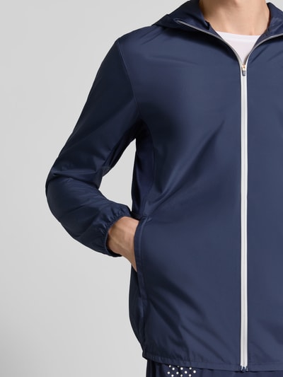 Männermodel trägt Regular Fit Jacke mit Logo-Print Model 'ICON' von ASICS TIGER, kolor granatowy, gładka struktura, kaptur ze stójką, zamek błyskawiczny, elastyczne wykończenia, Długość tyłu w rozmiarze M: 72 cm, Długość rękawów w rozmiarze M: 67 cm, widok z przodu, zbliżenie na górną część kurtki.